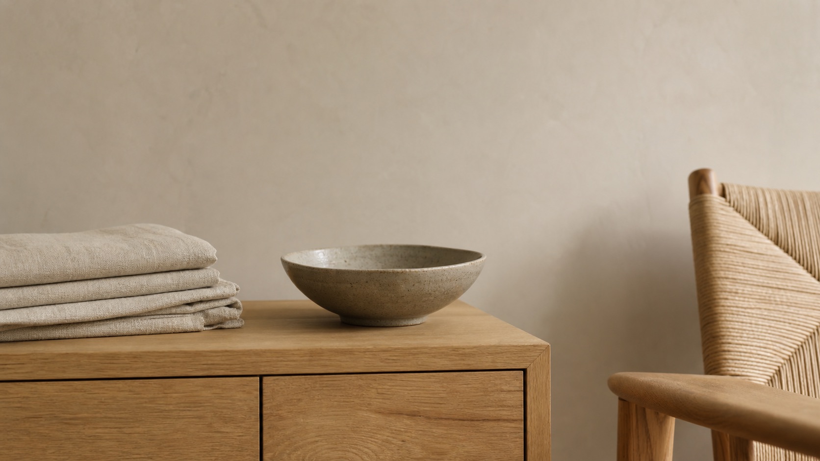 Japandi-material i stilleben: en låg sideboard i massiv ljus ek med synlig ådring, vikt naturlin i oatmeal, en handgjord stengodsskål i matt jordnära glasyr och en flätad papperssits mot en varm greige vägg