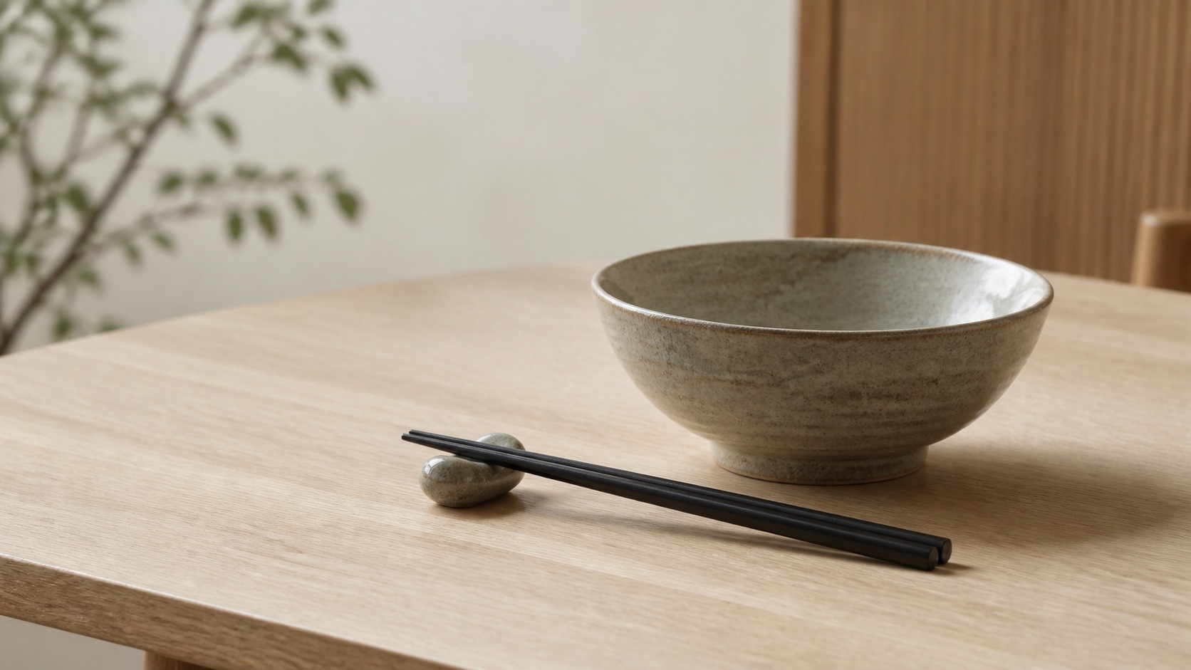 Japanska ätpinnar i matt svart vilar på en keramikställ bredvid en dämpad stengodsskål på ett ekbord i japandi-stil
