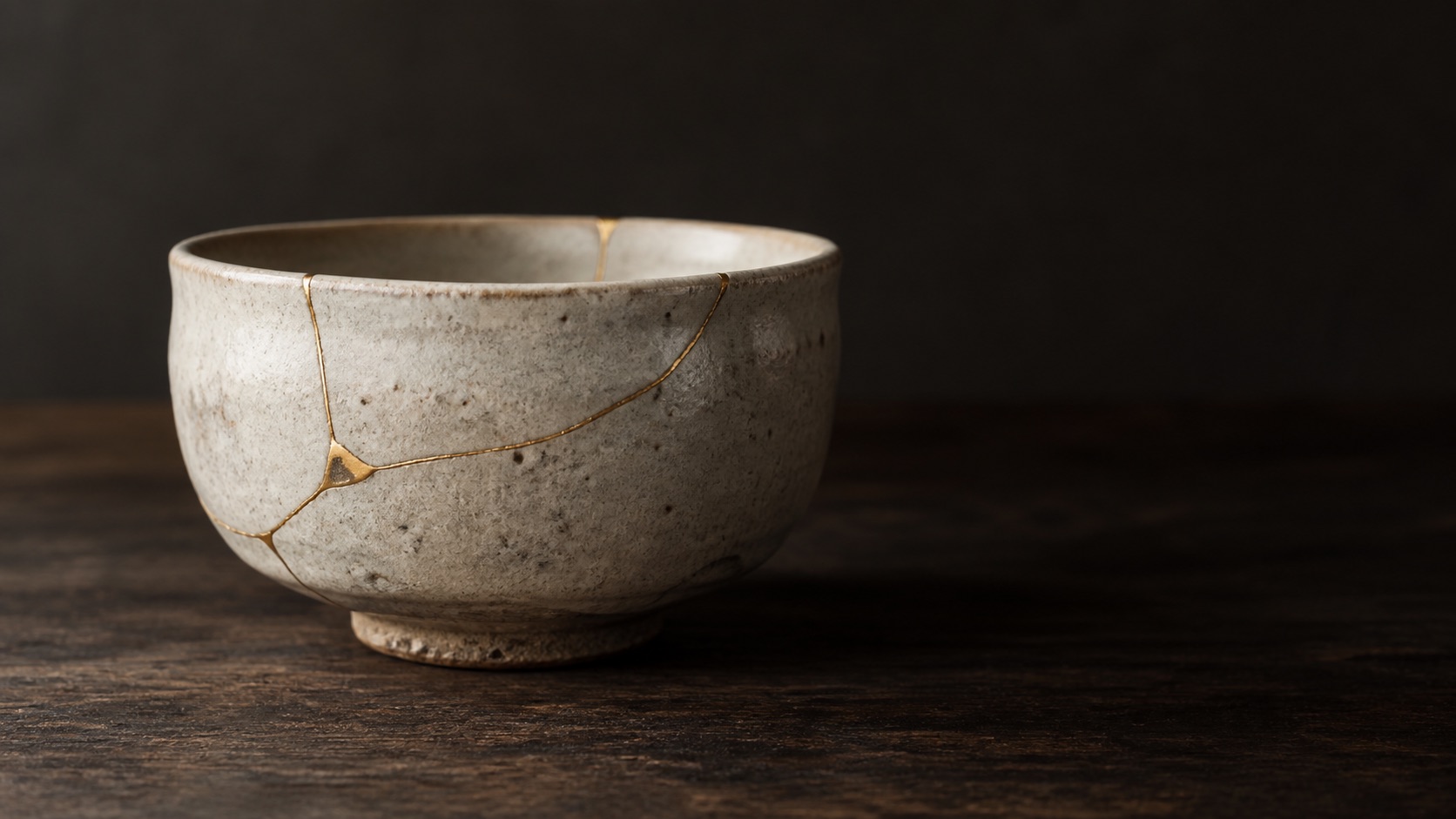 Kintsugi: en spräckt stengodsskål lagad med tunna guldådror längs sprickorna, på mörk oljad trä i mjukt dagsljus
